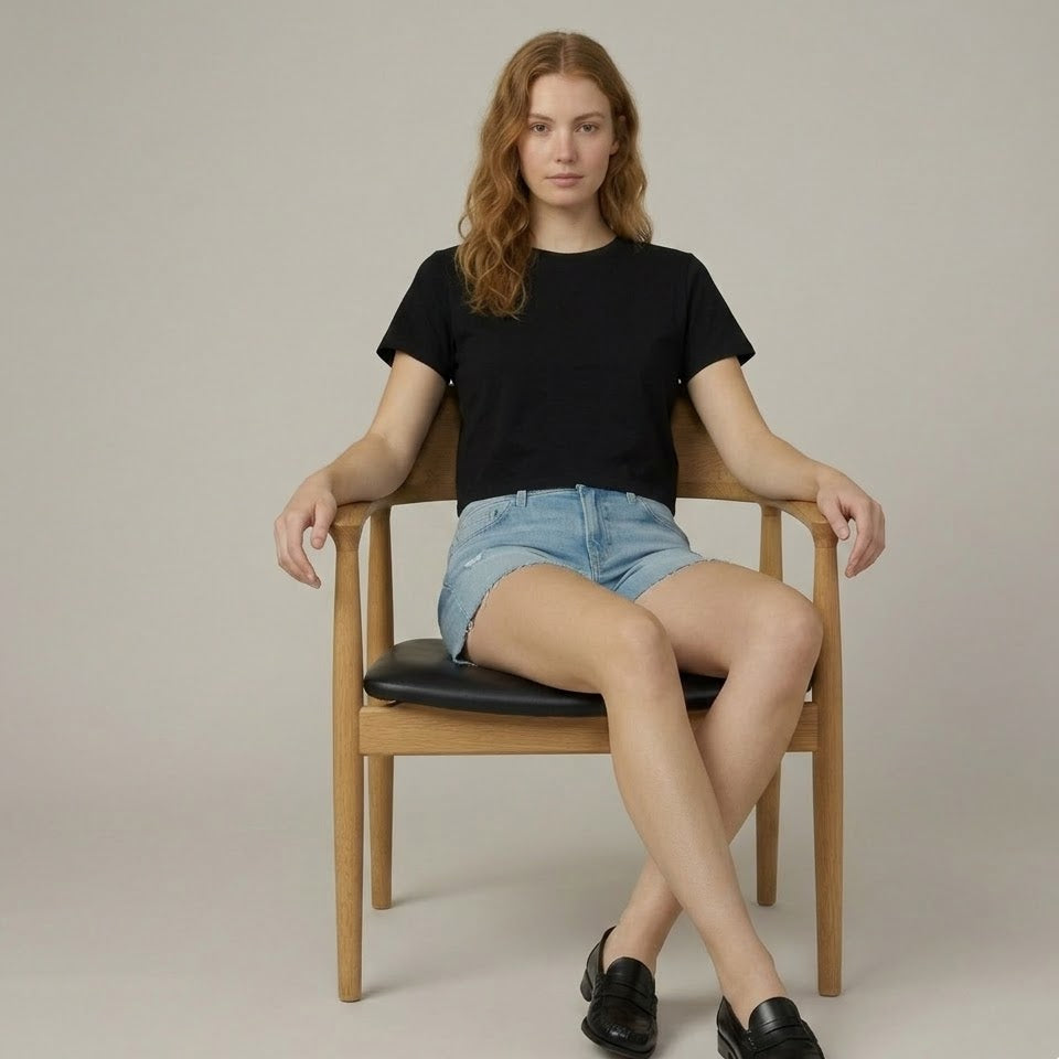 Woman sitting on a chair wearing a black t-shirt, denim shorts, and black shoes against a plain background
